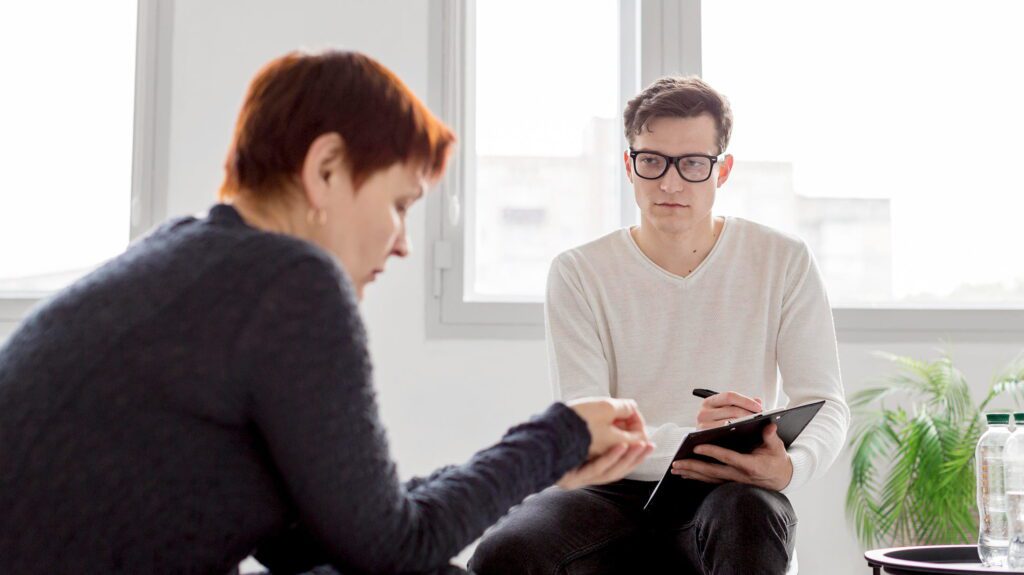 individual therapy session with female client and male therapist taking notes in bright counseling office with large windows and plants