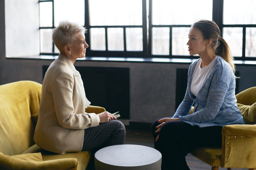Side view image of middle aged female psychologist discusses the problems with young pregnant woman, sitting in armchairs, facing each other.